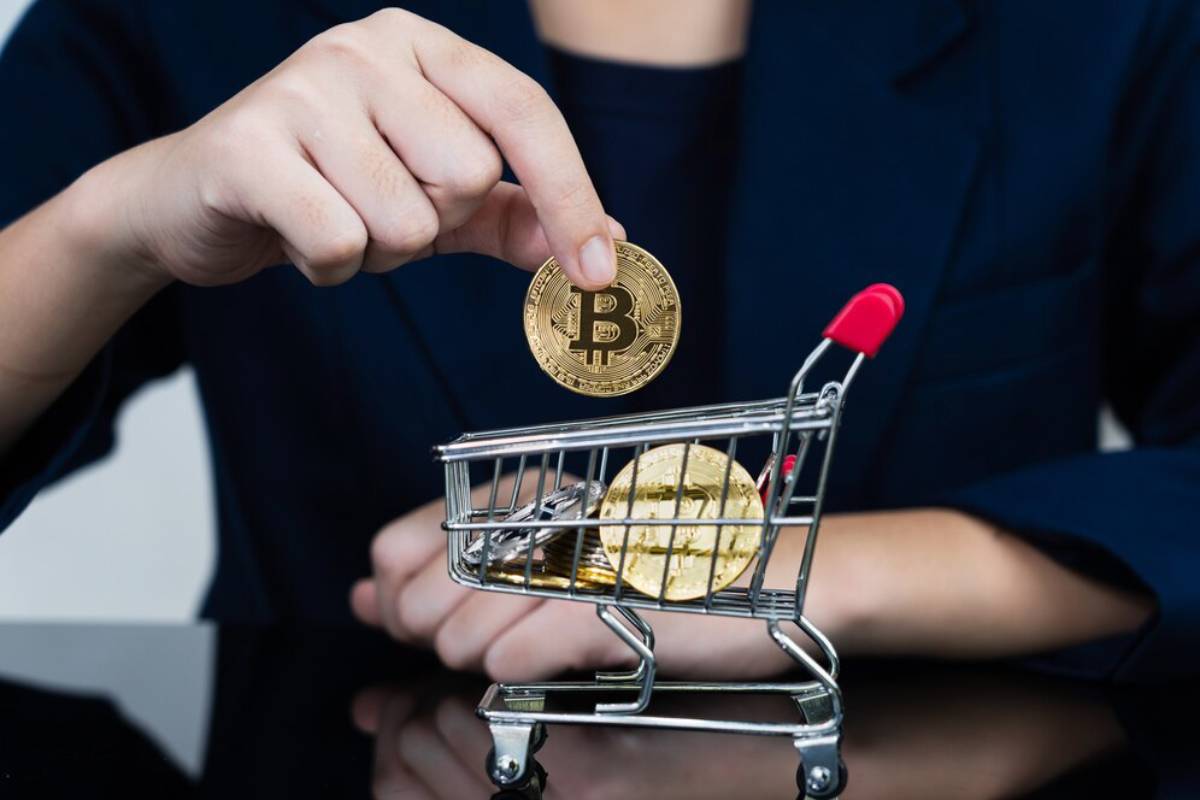 A person placing a Bitcoin coin into a miniature shopping cart, illustrating cryptocurrency transactions or investments.