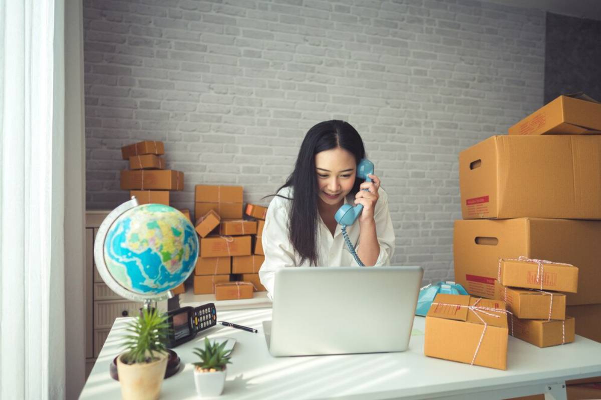 Woman talking on phone while using laptop, surrounded by packed boxes in an office setting.