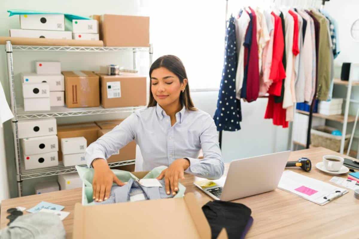 A person organizes clothing items at a desk with a laptop, surrounded by colorful garments and cardboard boxes.