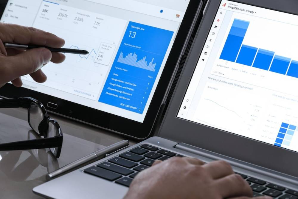 A person analyzes digital analytics data on a tablet and laptop, focusing on user statistics and trends with a pen in hand.