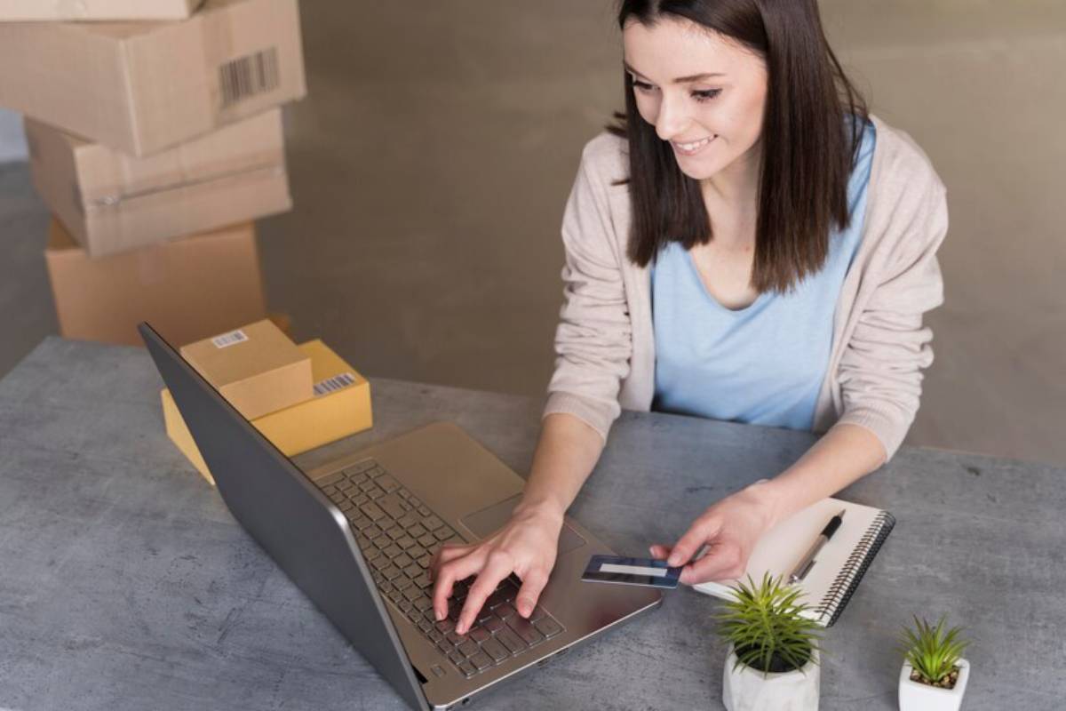 A person using a laptop while holding a credit card, with cardboard boxes stacked in the background and small plants on the desk.