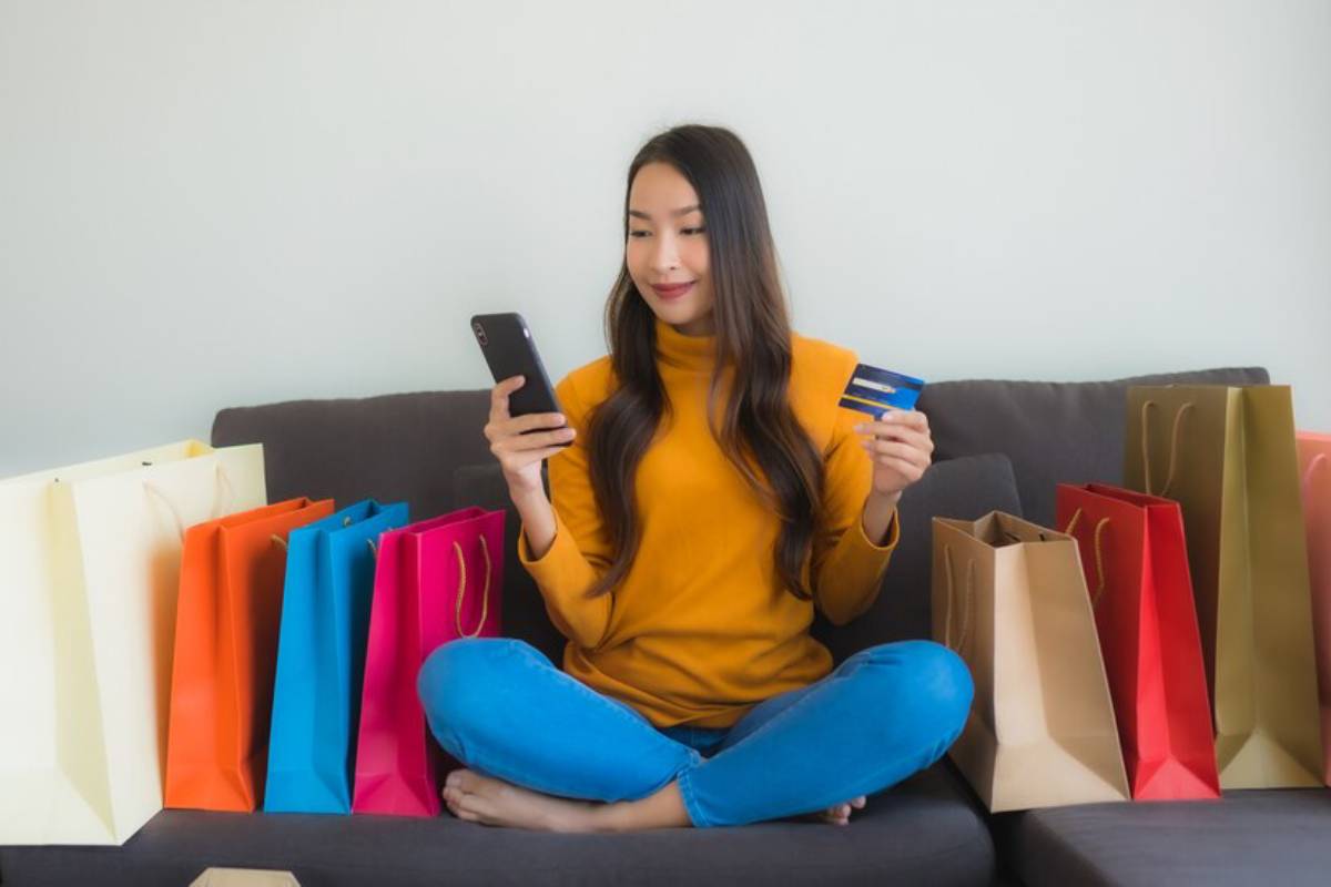 A person in a yellow sweater sits cross-legged on a couch, holding a phone and a credit card, surrounded by colorful shopping bags.