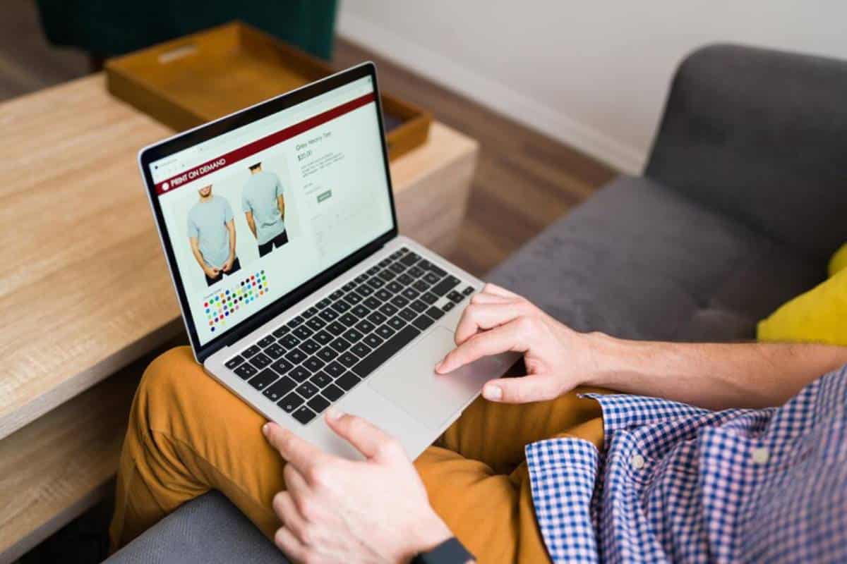 A person sits on a couch browsing a laptop, viewing a t-shirt product page.