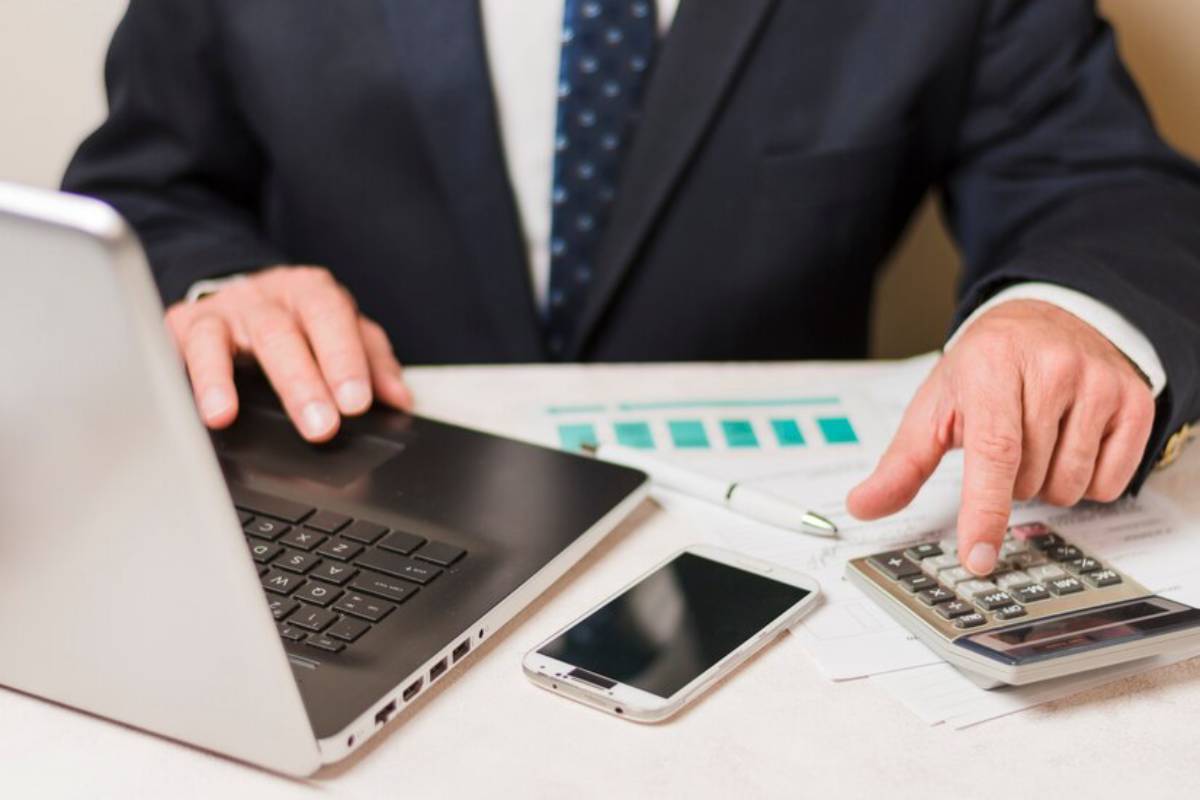 A businessman in a suit using a laptop and calculator, with a smartphone and documents on a desk.