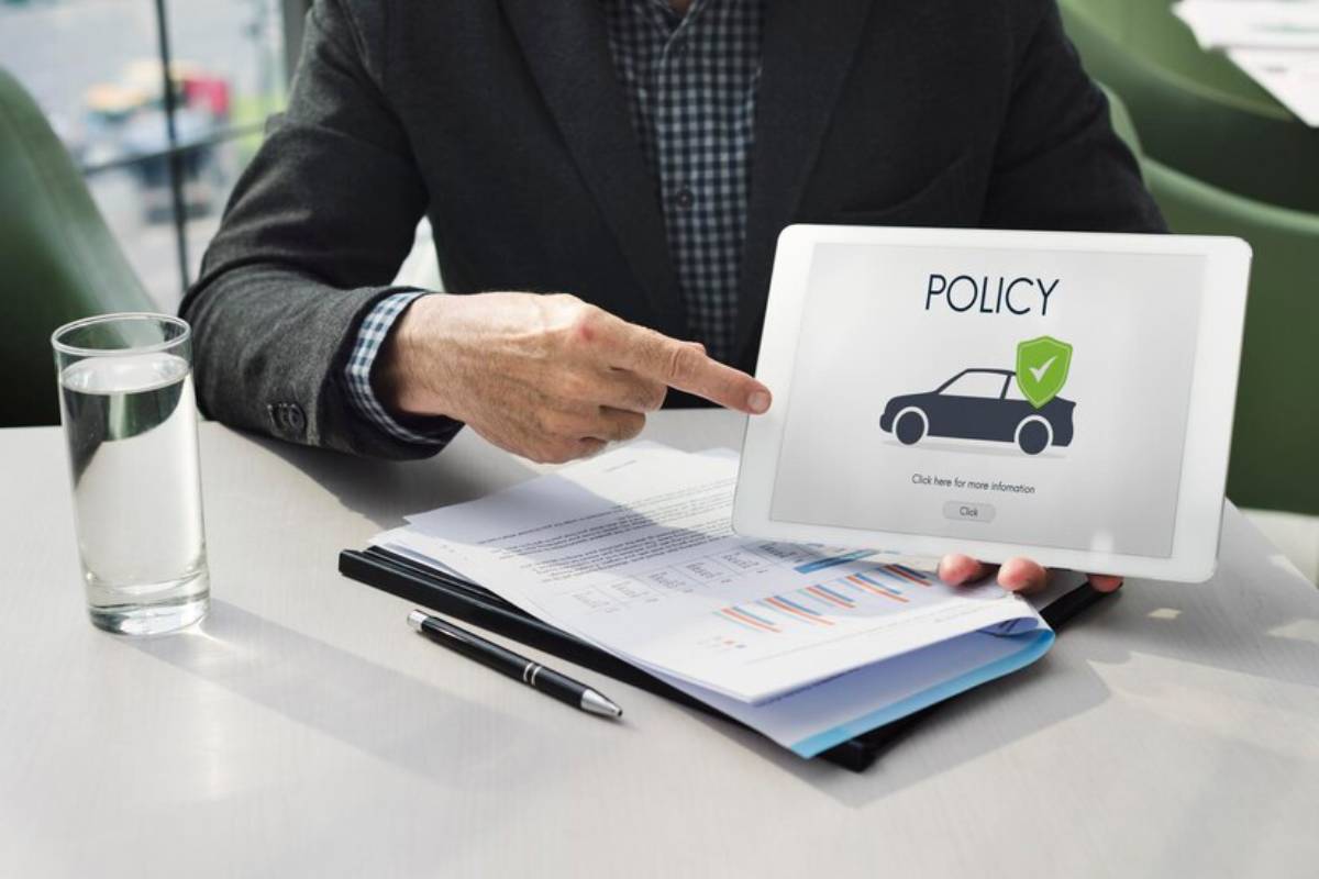 A person in a suit points at a tablet displaying a insurance policy, with a glass of water and documents on the table.