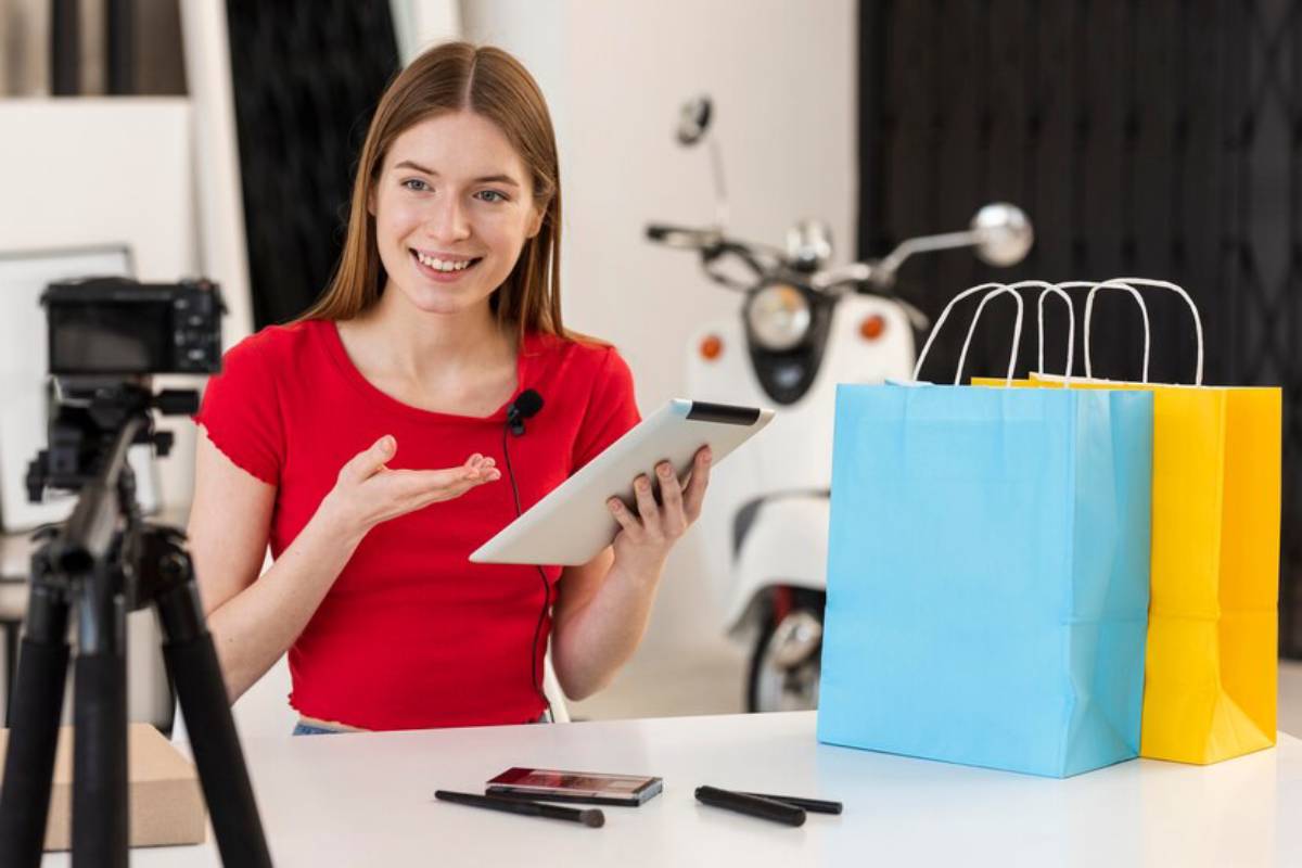 A person in a red shirt is recording on camera while holding a tablet, with shopping bags in blue and yellow on the table beside them.