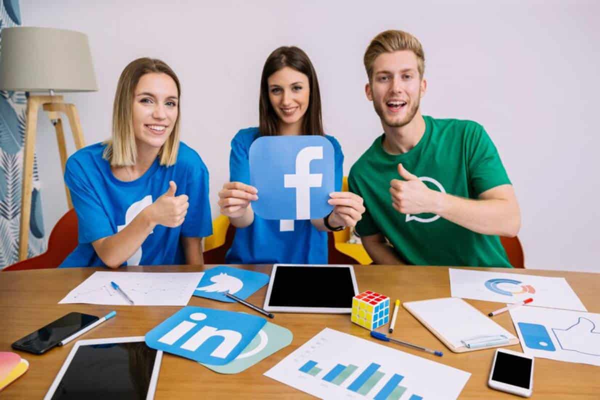 Three individuals in casual attire display a Facebook logo and thumbs-up signs, with marketing materials and devices on a table.