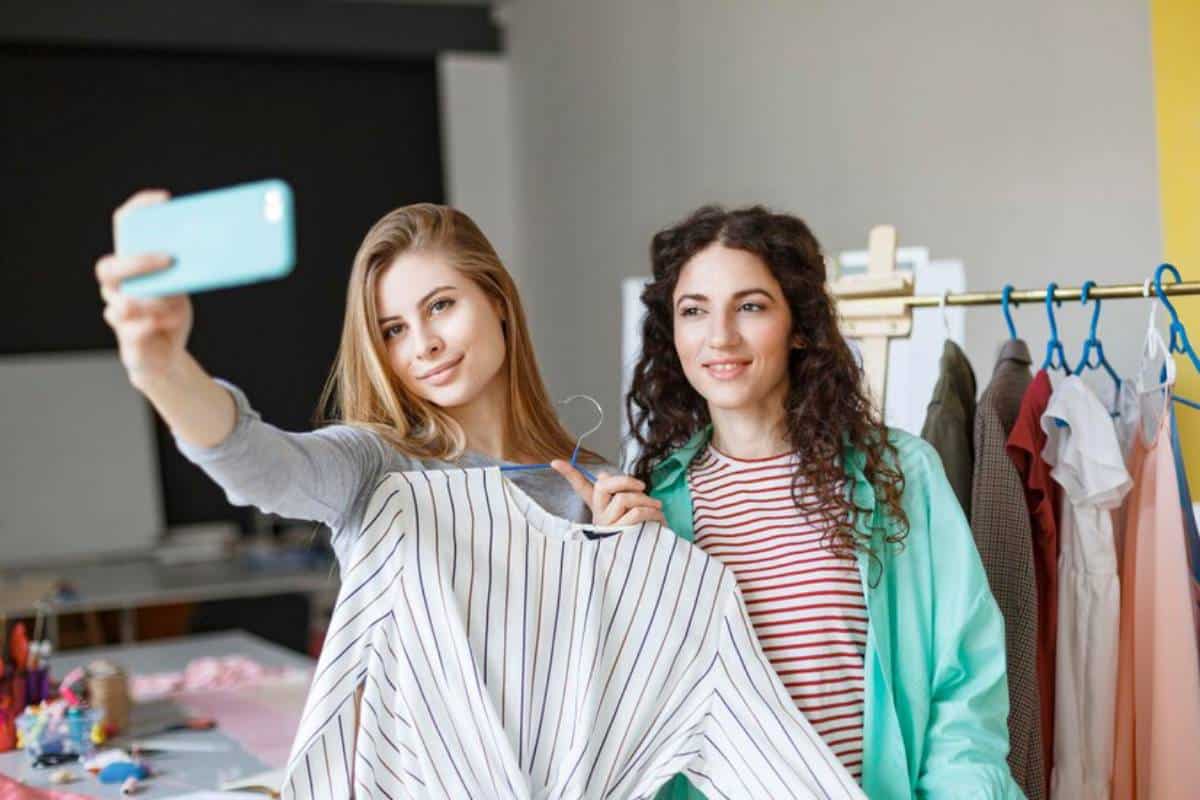 Two young women recording on phone, one holding a striped shirt, with colorful clothing displayed on a rack in the background.