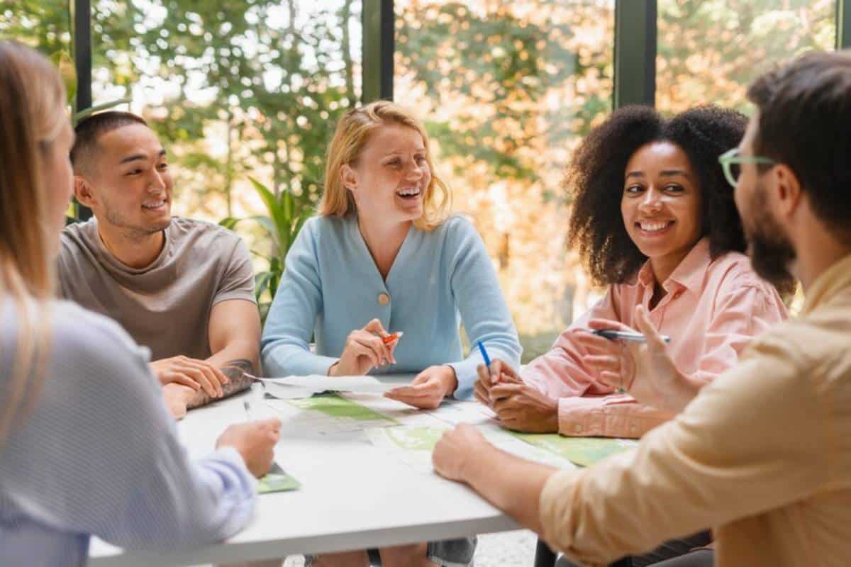 A diverse group collaborates around a table covered with maps, engaged in discussion, with greenery in the background.