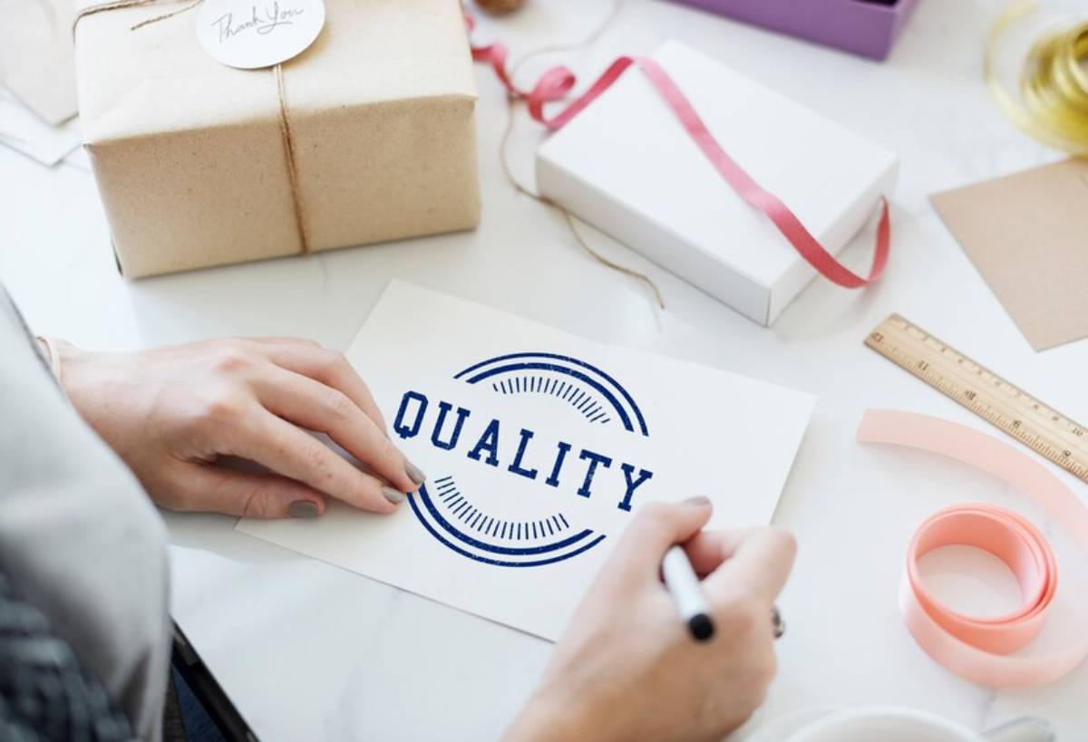 A person writes "QUALITY" on a card, surrounded by gift-wrapping supplies, a ruler, and wrapped presents on a table.