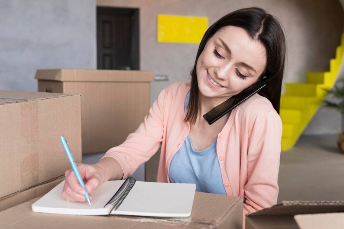 A person in a pink cardigan sits amidst cardboard boxes, writing notes and talking on call.