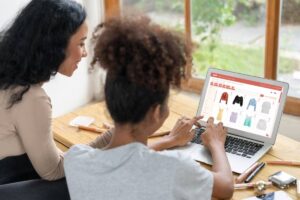 Two women shopping online on a laptop, viewing a clothing website by a window.
